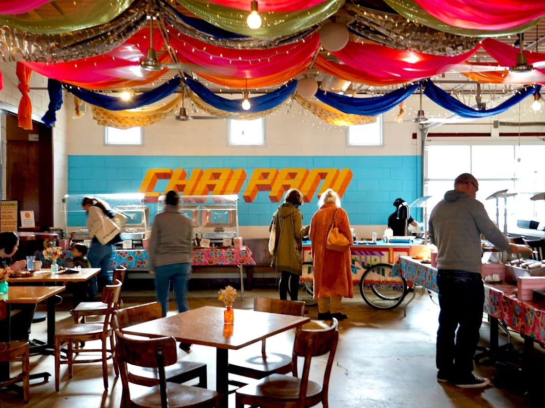 The interior of Chai Pani, a restaurant with colorful draped fabrics on the ceiling and a buffet line, with several patrons.
