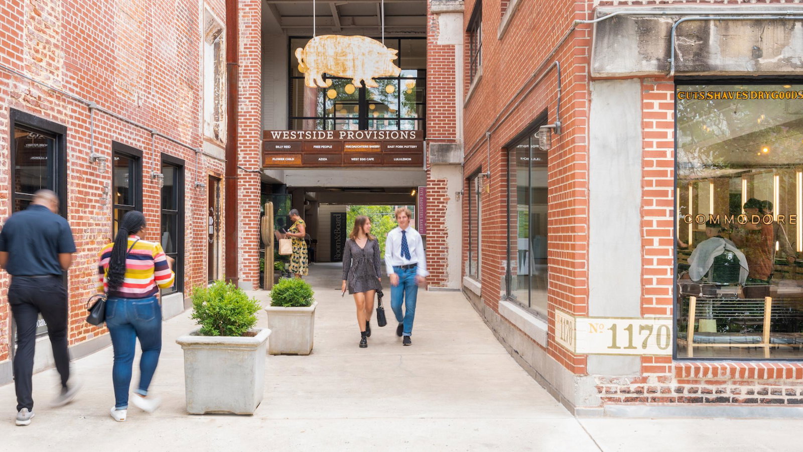 Pedestrians walk through the open-air brick promenade at Westside Provisions District in Atlanta, with a large golden pig sculpture suspended above a directory sign listing tenants including West Egg Cafe and Free People. The storefront at No. 1170 houses Commodore, a cuts and drygoods shop.