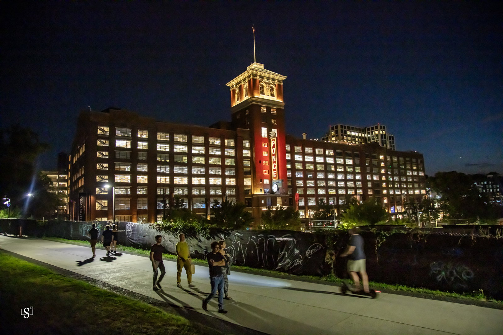 Ponce City Market, a massive illuminated red-brick building with a gold clock tower and vertical red neon PONCE sign, seen at night from the BeltLine Eastside Trail with pedestrians walking in the foreground.