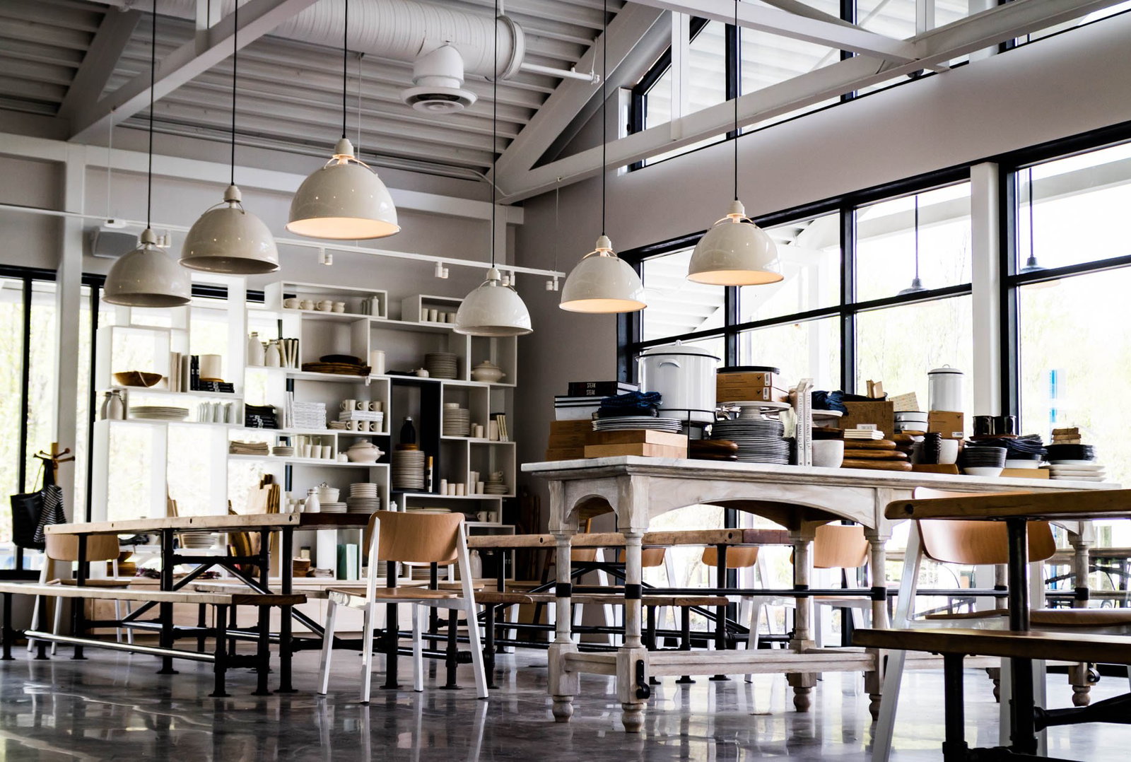 A spacious, modern interior with high ceilings, large windows, and multiple white pendant lights hanging above wooden tables and chairs. Shelves