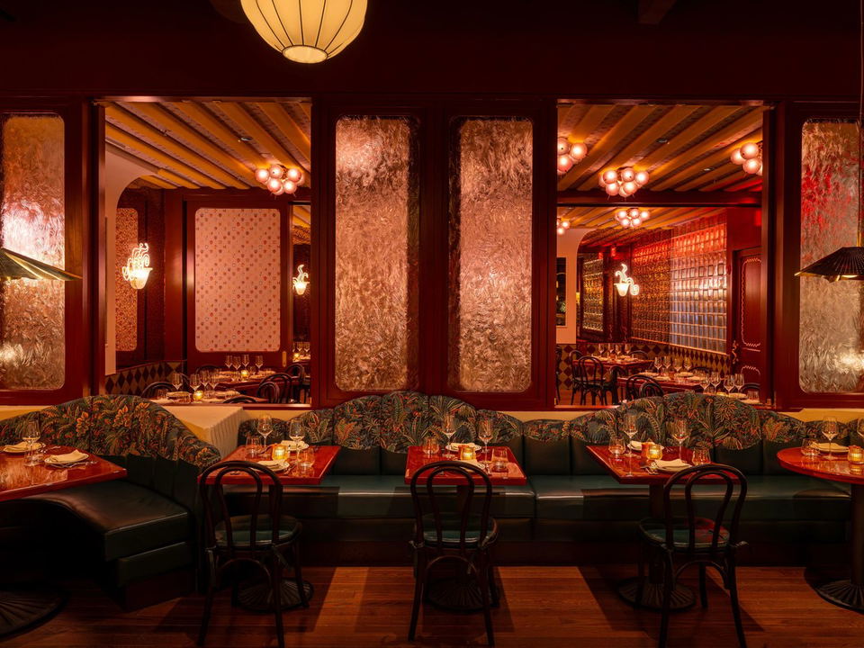 A warmly lit, maximalist Italian-American restaurant dining room, featuring tropical-print banquette seating and red lacquered tables.