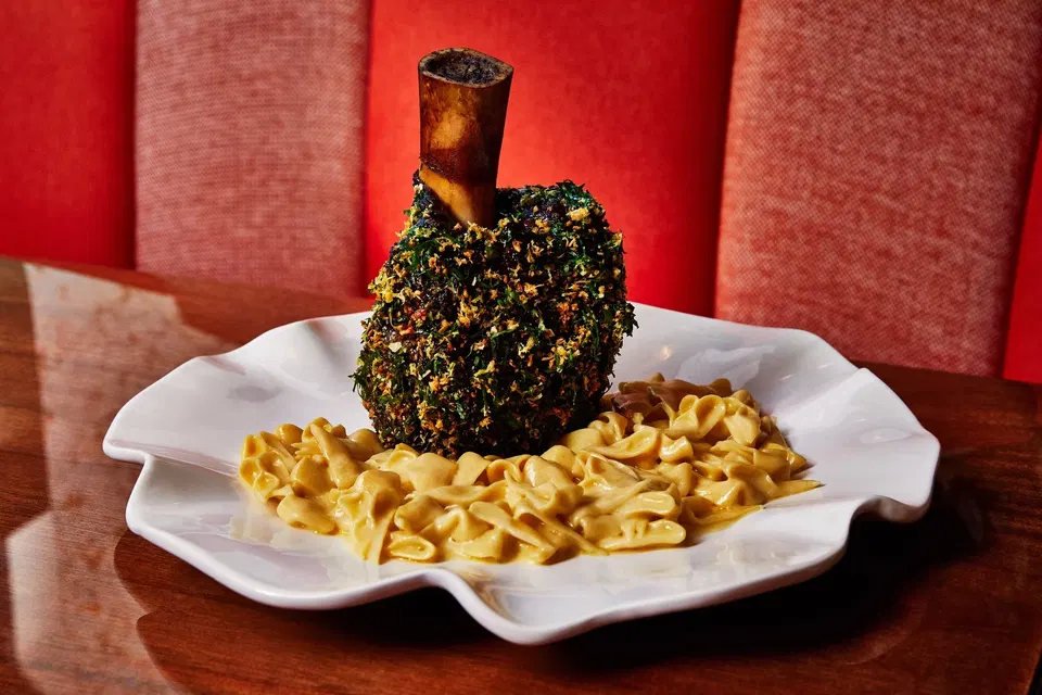 A herb-crusted veal shank osso buco with exposed bone stands upright on a white ruffled plate, surrounded by creamy pasta, set against a red banquette.