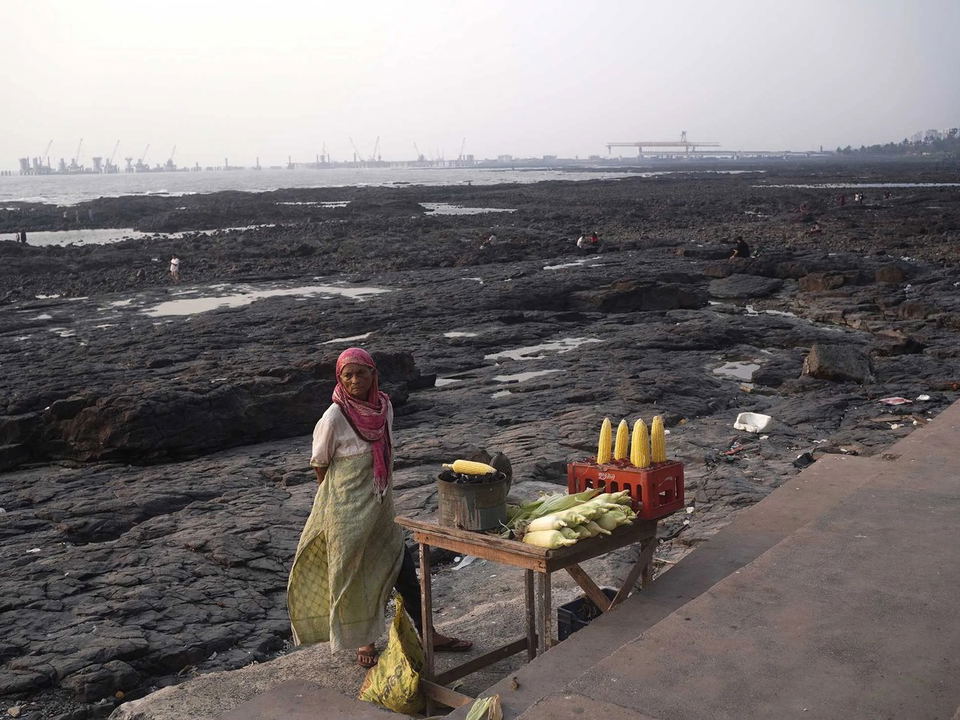 Bandra's rocky shoreline, with a woman selling corn and industrial infrastructure in the distance.