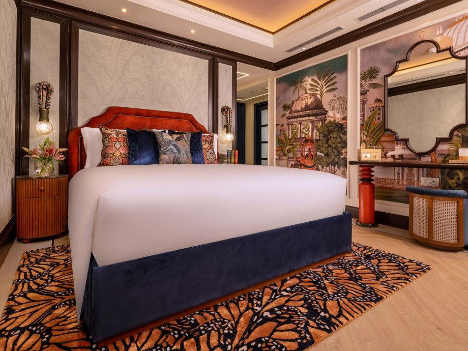 A luxurious hotel guestroom with a king bed, rust-red velvet headboard, navy bed skirt, patterned pillows, and a monarch butterfly rug.