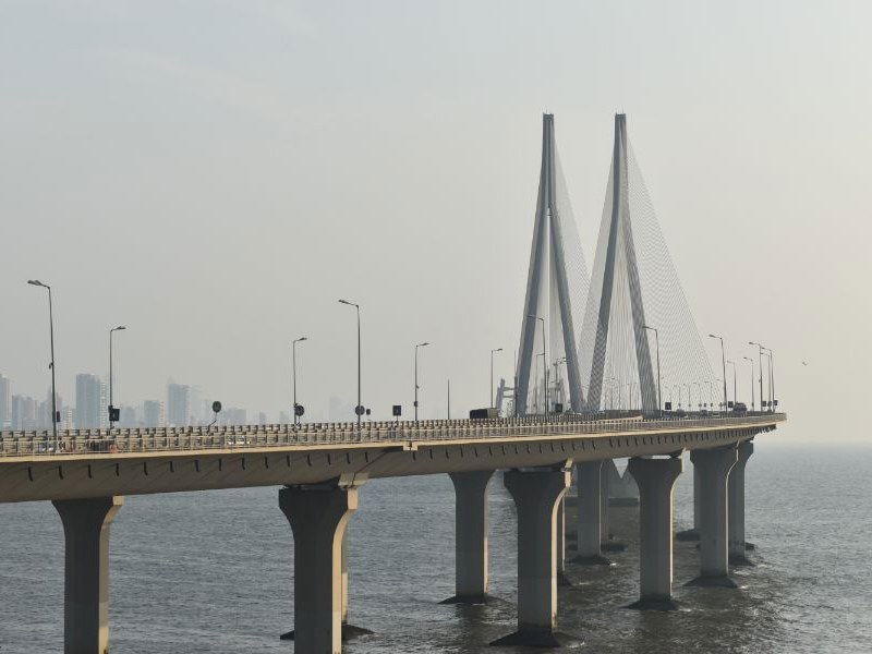 A long, elevated cable-stayed bridge, the Bandra-Worli Sea Link, stretches over the sea with a hazy city skyline in the background.