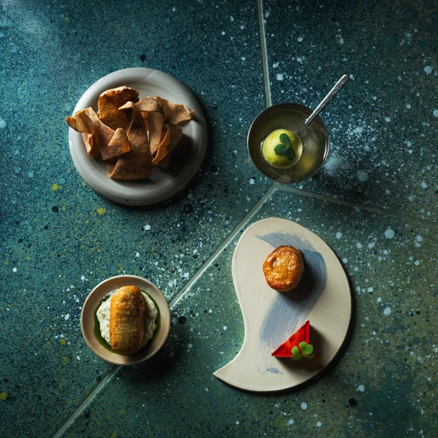 High-end plated bar snacks and cocktail components arranged on ceramic plates on a teal surface, showing fine-dining presentation style consistent wit