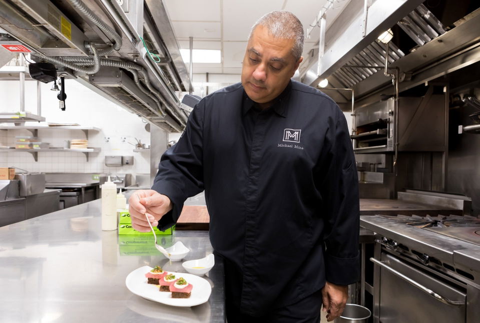 A chef in a black chef's coat with 'Michael Mina' embroidered on it is plating a dish in a professional kitchen; the source page (Eater SF) explicitly