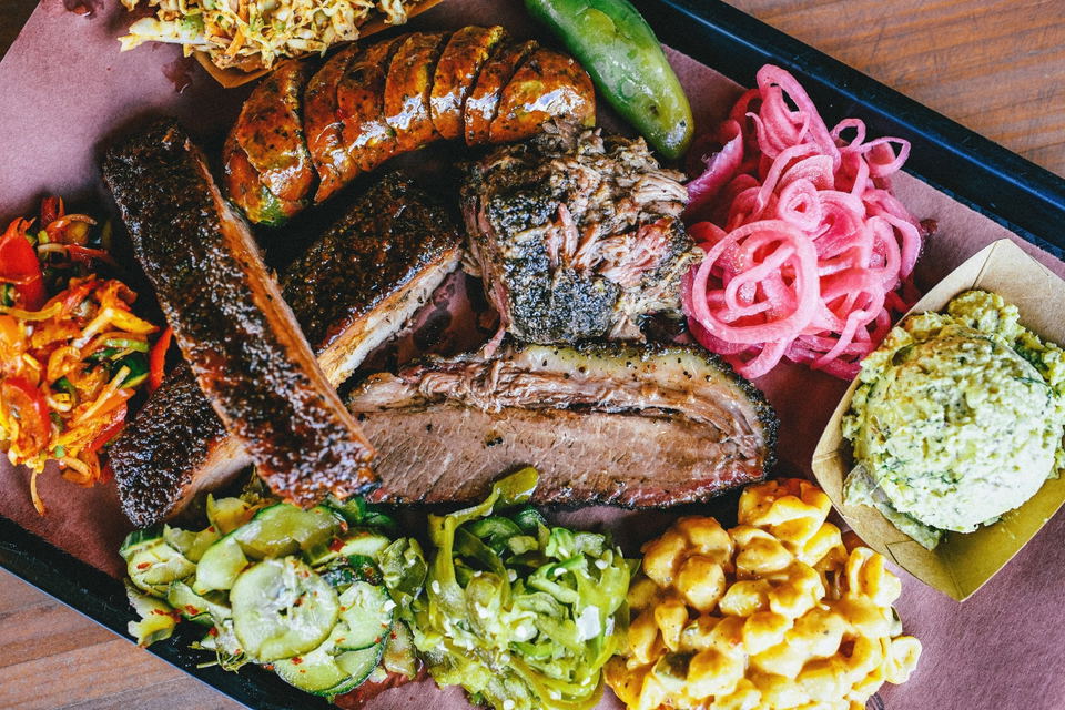 A plated meat and vegetable board with grilled steak slices, sausage, pickled red onions, grilled cucumber, corn, and herb-topped spreads on a dark plate.