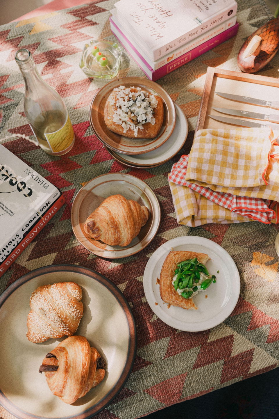 Overhead view of a brunch table with baked goods on plates, a bottle of wine, stacked books, and checkered napkins on a geometric patterned blanket.
