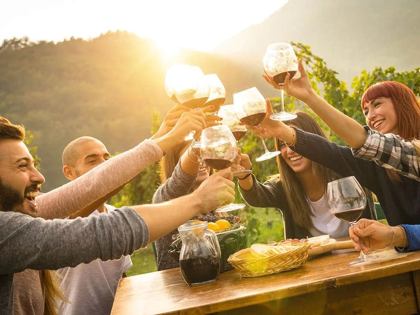 Five people raising wine glasses in a toast at a wooden table with food and wine bottles, with vineyard hills and sunset in the background.