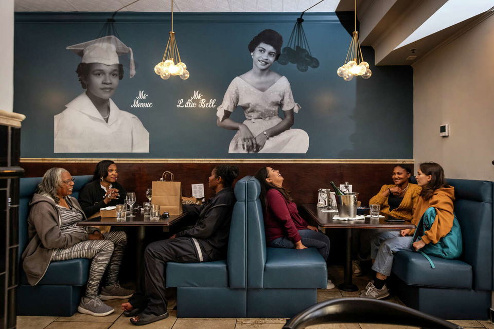 Six women are seated at tables in Minnie Bell's Soul Movement, with large black and white portraits of women on the wall.