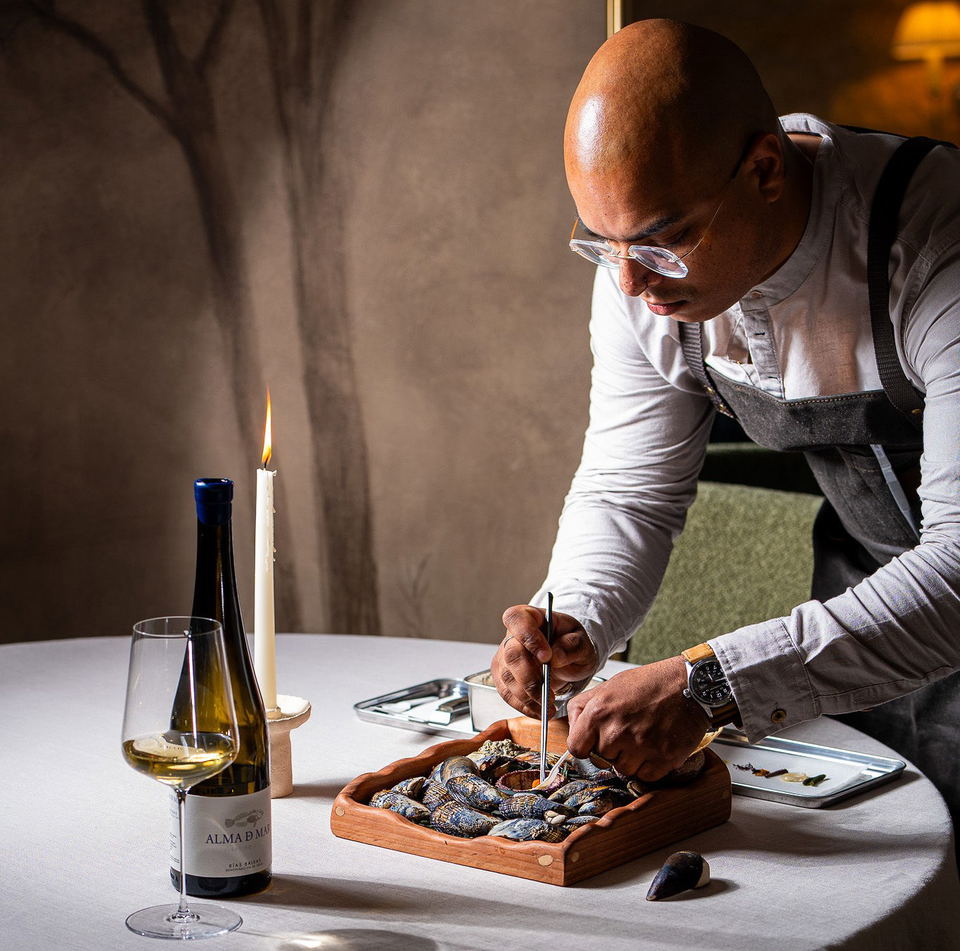A chef in a white shirt and dark apron meticulously prepares a dish of mussels on a wooden tray, with a bottle of Alma D Mar wine and a lit candle on