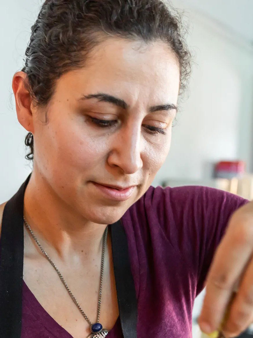 Reem Assil, a woman with curly hair in a purple shirt and black apron, drizzles honey onto a knafeh sandwich.
