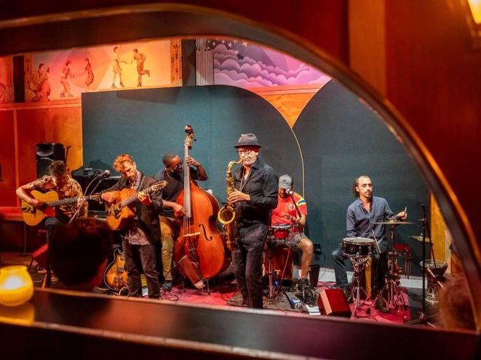 The DeLuxe: A jazz band performs on a warmly lit Art Deco stage with painted murals behind them.
