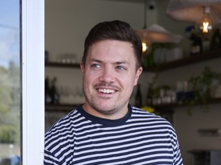 A man with dark hair and a mustache smiles at the camera while wearing a striped shirt indoors.
