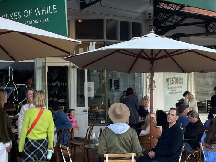 Patrons gather at outdoor tables under large umbrellas in front of a building with 'WINES OF WHILE' and 'WESTONS BARBERSHOP' signs.