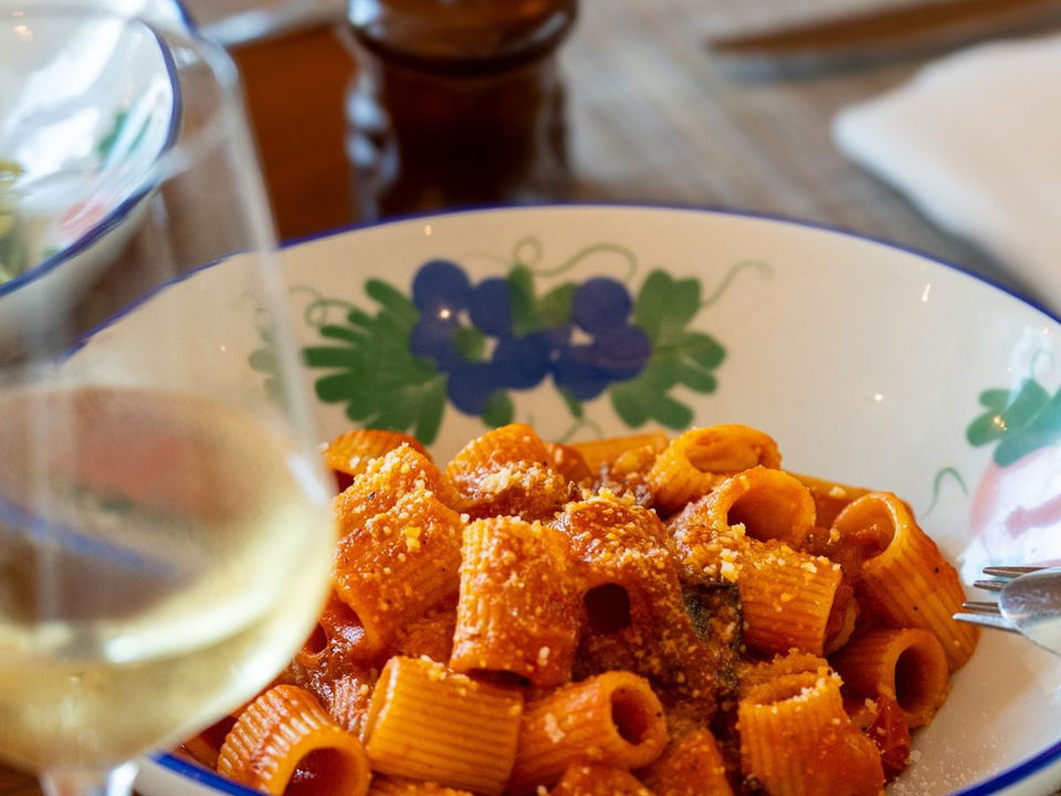 A close-up of a bowl of rigatoni pasta in a rich red tomato sauce, topped with grated cheese, served on a white table with a glass of white wine.