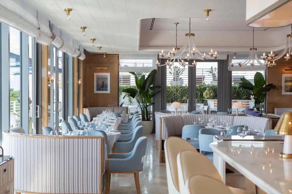 The bright, airy dining room of Bellini Restaurant Coconut Grove, featuring light blue chairs, striped banquettes, and large windows overlooking a