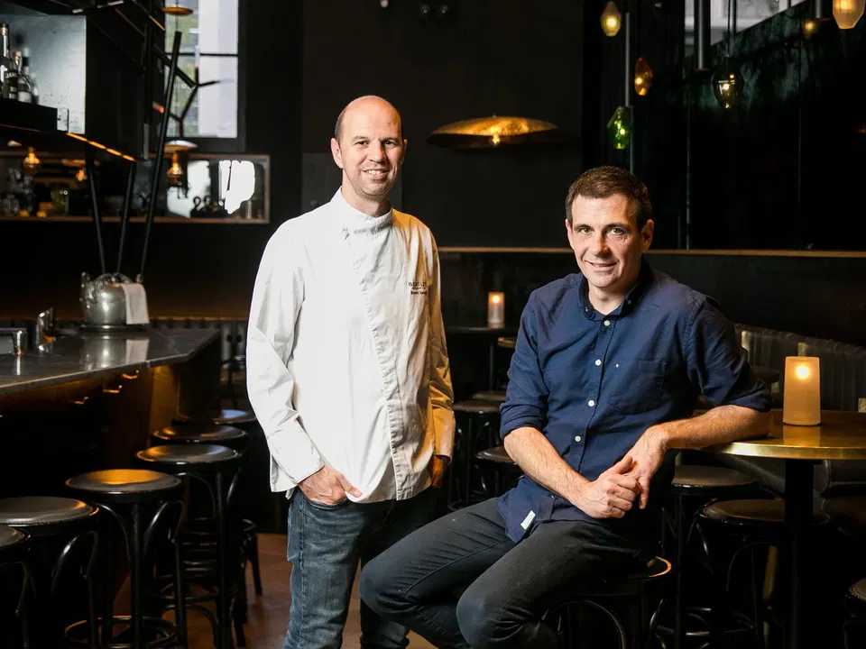 Chef Brent Savage in a white Bentley-embroidered chef's coat stands alongside sommelier Nick Hildebrandt in a dark blue shirt, both smiling in the moody, candlelit interior of Bentley Restaurant + Bar, Sydney, surrounded by black bar stools, brass pendant lights and dark timber finishes.