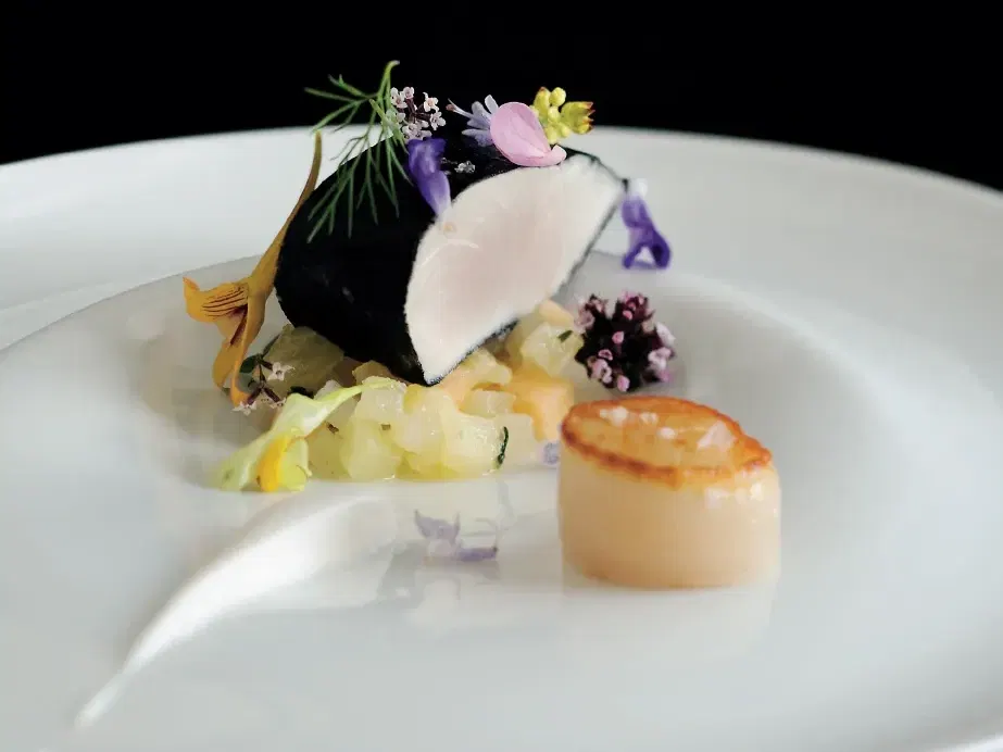 A fine-dining plate featuring a portion of squid-ink-marinated kingfish topped with a thin slice of white fish and edible flowers in purple, pink, and yellow, alongside a golden seared scallop and a bed of diced pale fruit, served on a white plate against a black background.