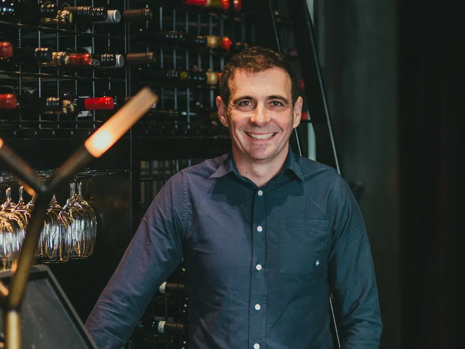 A smiling middle-aged man in a dark blue shirt stands in front of an extensive wall-mounted wine rack filled with bottles.