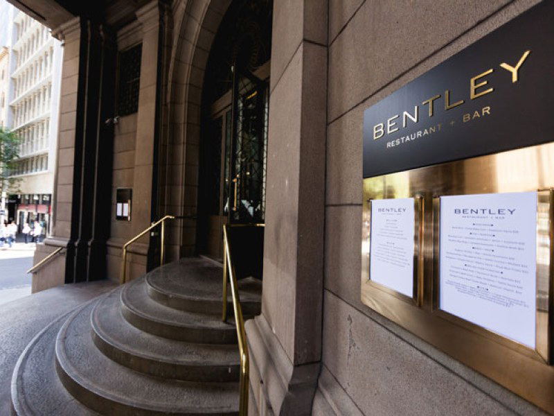 The exterior of Bentley Restaurant + Bar, featuring a black sign with gold lettering, curved stone steps, and brass railings leading to an arched