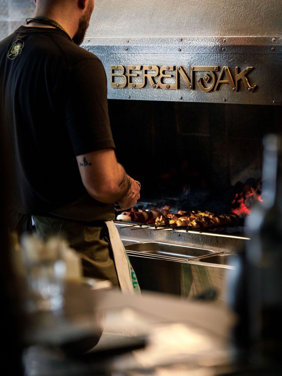 A man in a dark shirt stands at a grill with the Berenjak nameplate, cooking meat over hot coals and flames.