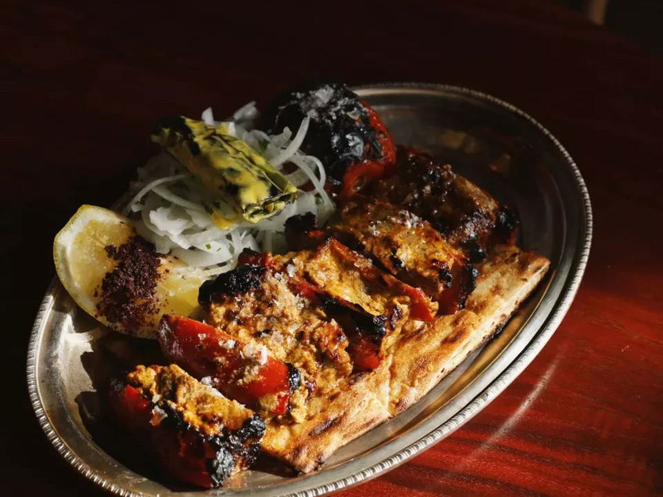 A metal plate containing grilled meat kebabs with char marks, accompanied by grilled tomato, sliced onions, fresh herbs, and a lemon wedge.