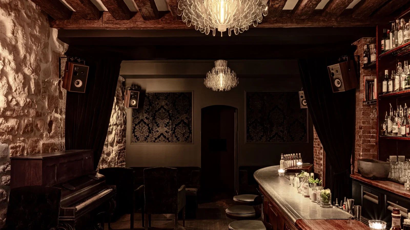 A dimly lit cocktail bar with a long, curved bar counter, stools, and shelves of liquor bottles. Chandeliers hang from the ceiling.