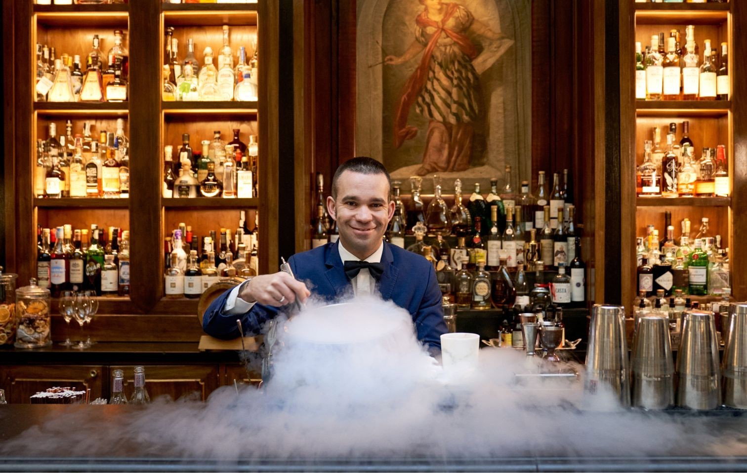 A smiling bartender in a blue suit and bow tie stands behind a bar, preparing a cocktail with a dramatic cloud of smoke.