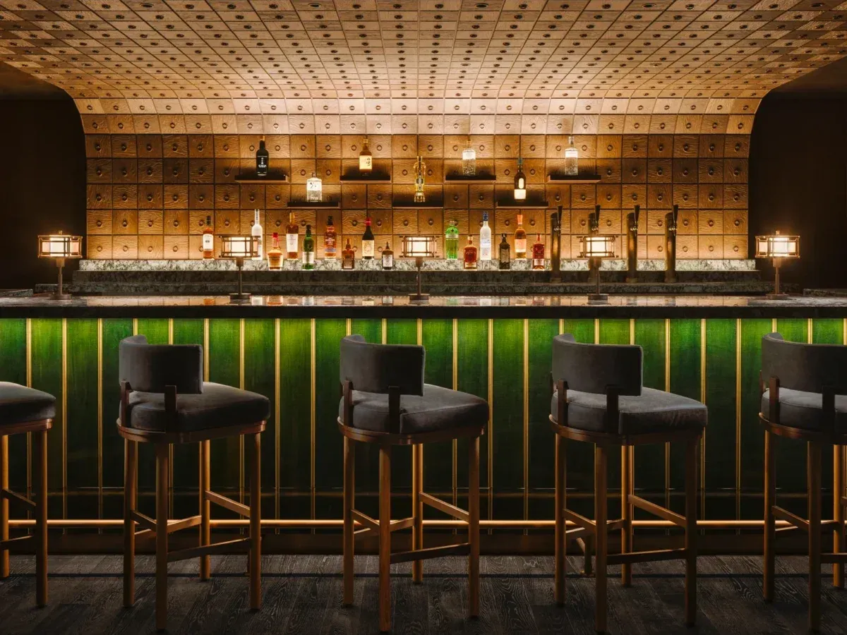 A dimly lit bar interior with a long, dark green counter and several high-backed bar stools. Behind the counter, shelves display numerous bottles.