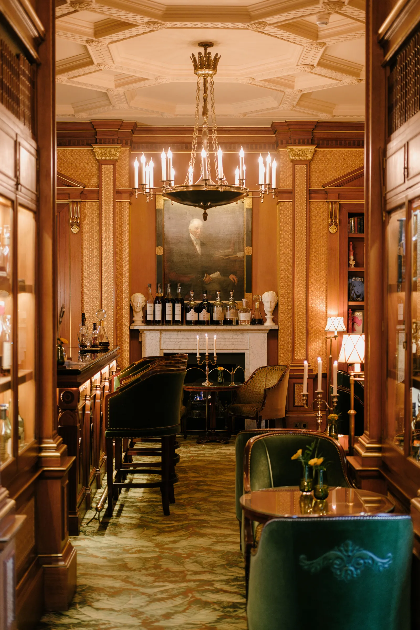 A luxurious cocktail bar with dark wood paneling, ornate gold accents, and a patterned ceiling, featuring a large chandelier.