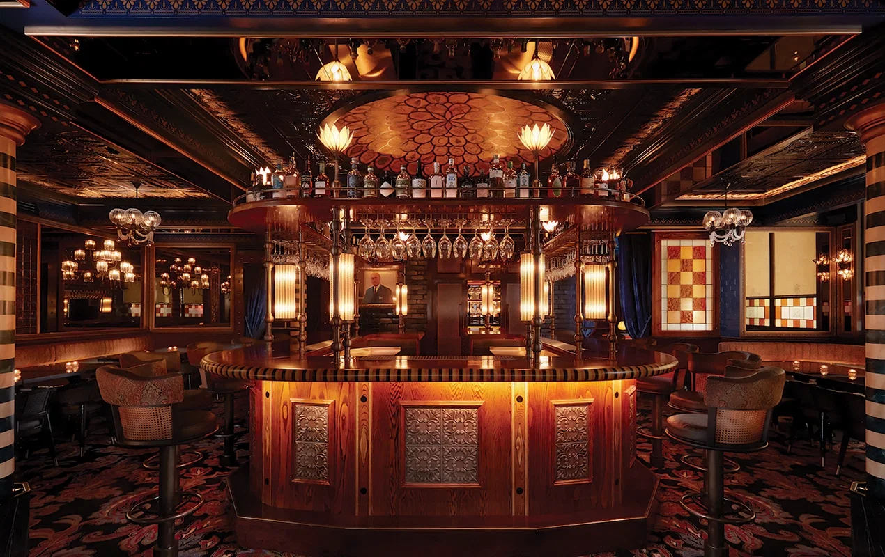 A dimly lit, opulent cocktail bar with a central, circular wooden bar, surrounded by high stools. Bottles line shelves above the bar, and chandeliers.
