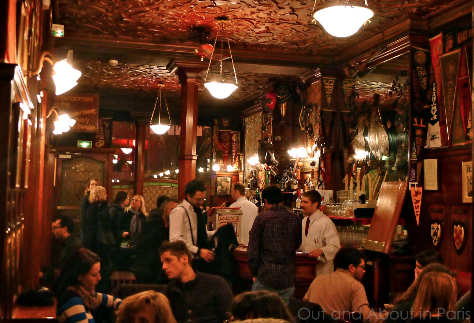 A dimly lit, ornate bar interior with a patterned ceiling, chandeliers, and wall sconces, filled with patrons and bartenders.
