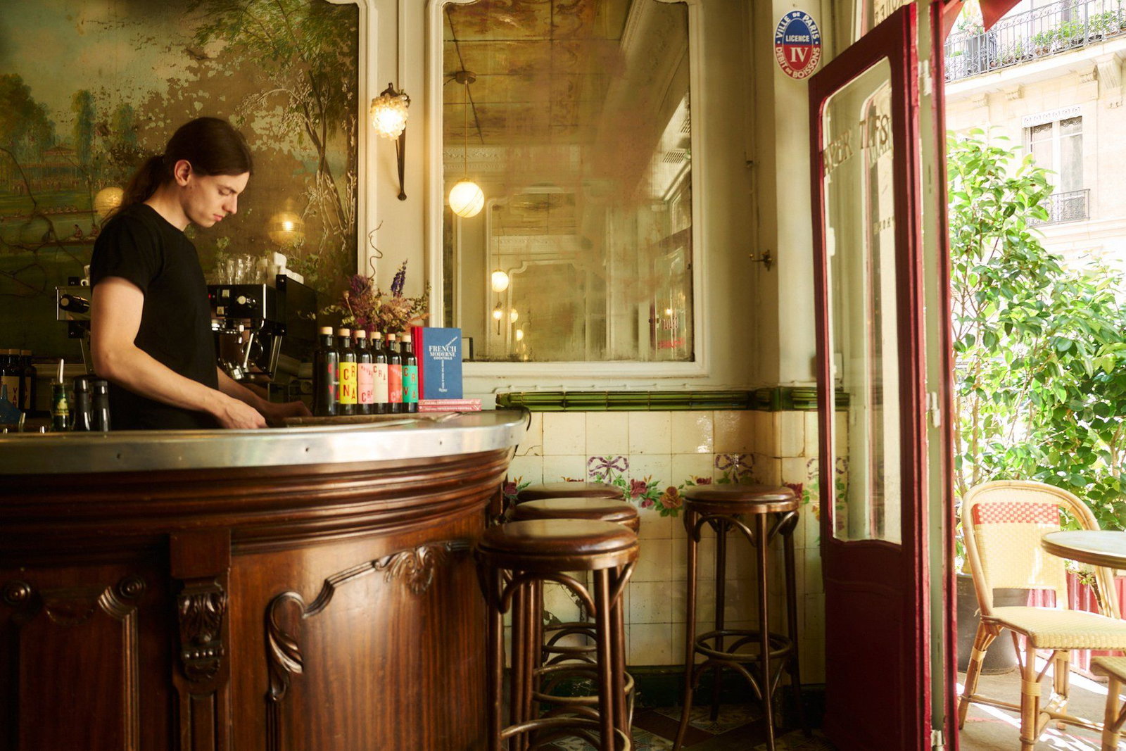 A bartender with long hair in a black t-shirt prepares drinks behind a wooden bar in a Parisian cocktail bar.