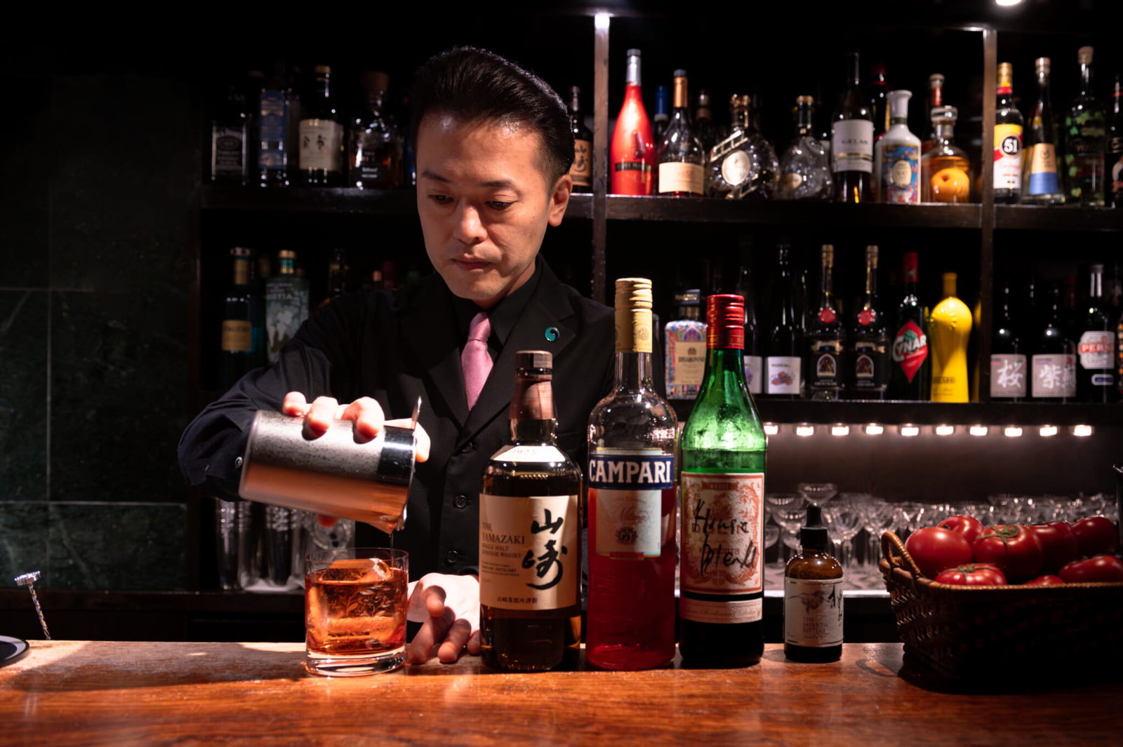 A bartender in a black suit and pink tie pours a drink from a metal shaker into a glass with ice. Bottles of Yamazaki, Campari, and other spirits are