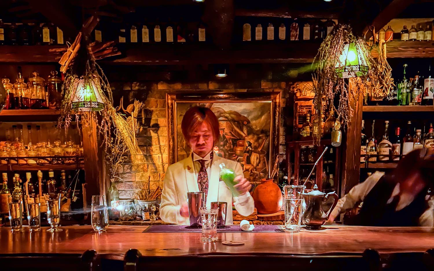 A bartender in a white suit and patterned tie pours a green liquid from a small bottle into a shaker at a dimly lit bar.