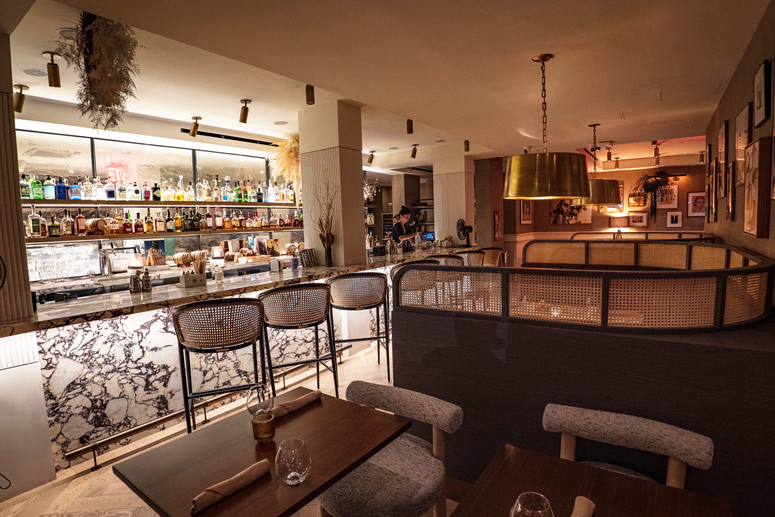 A well-lit bar interior with a marble counter, numerous liquor bottles on shelves, and seating areas with woven cane details.