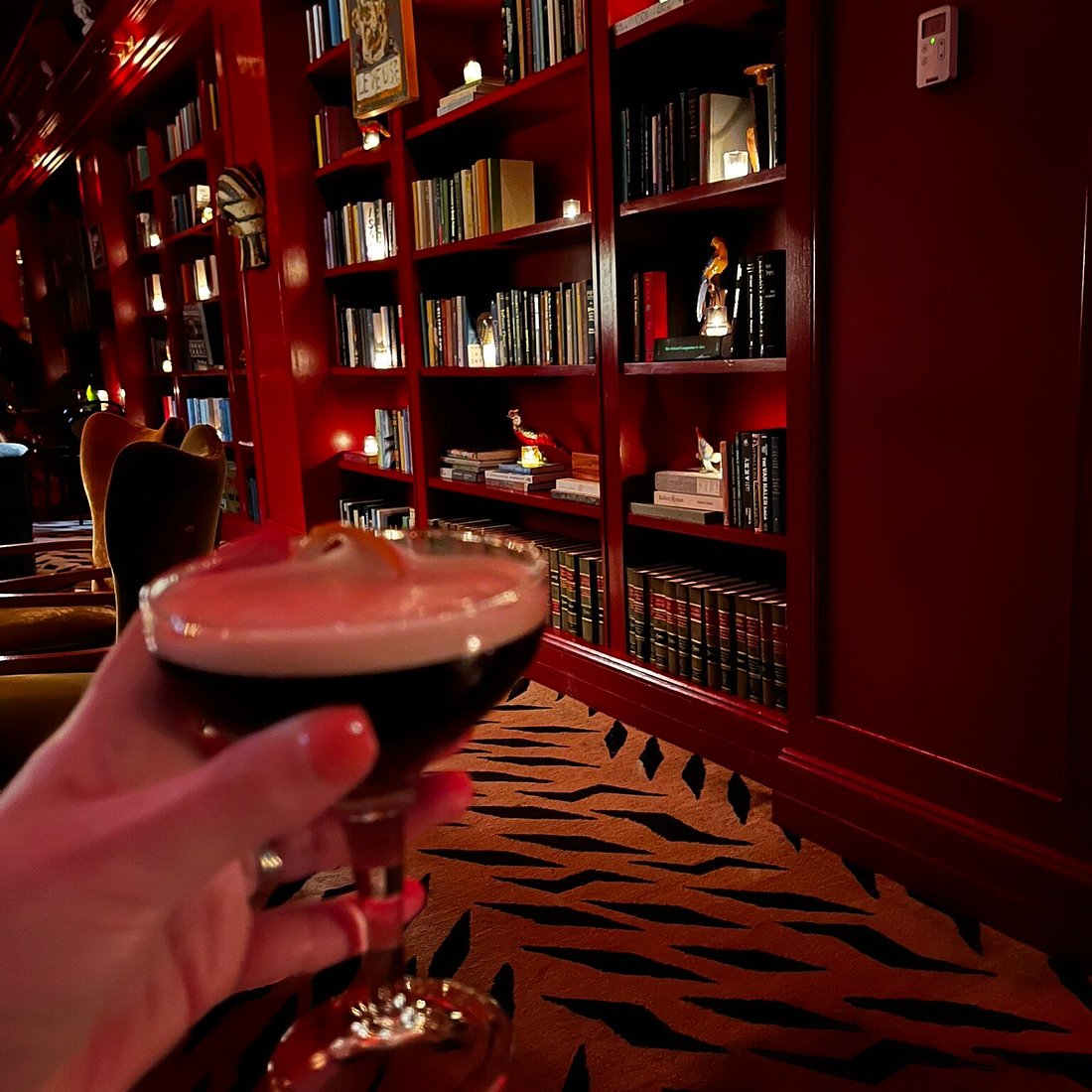 A hand holds a dark cocktail with a foamy top and orange peel garnish in a coupe glass, set against a backdrop of red bookshelves filled with books