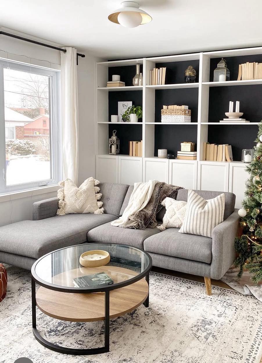 A modern living room features a grey sectional, glass coffee table, black-backed shelves, and a festive tree by a snowy window.