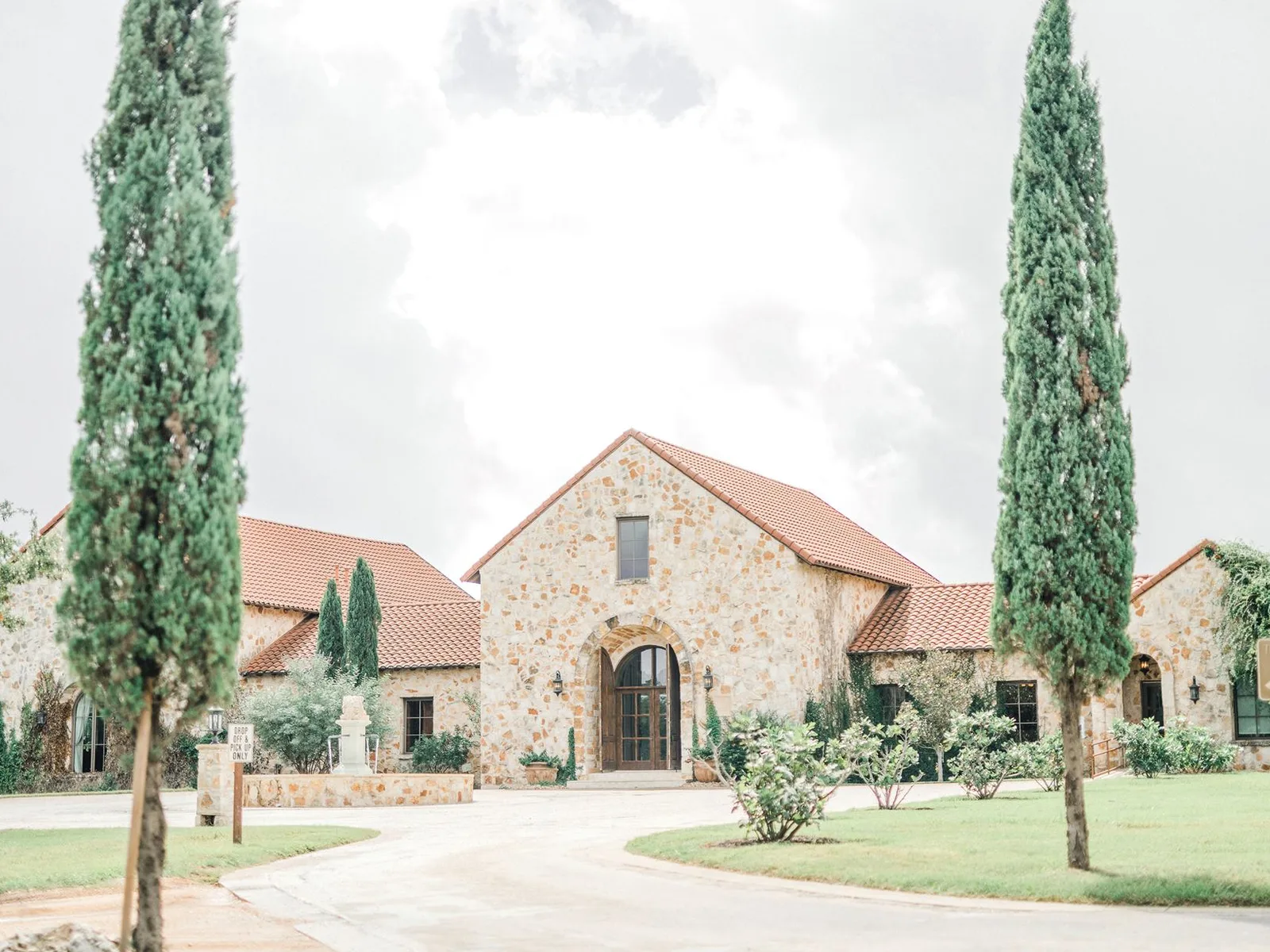 A large, light-colored stone building with a terracotta tile roof, featuring an arched entrance and tall, slender green trees on either side of a