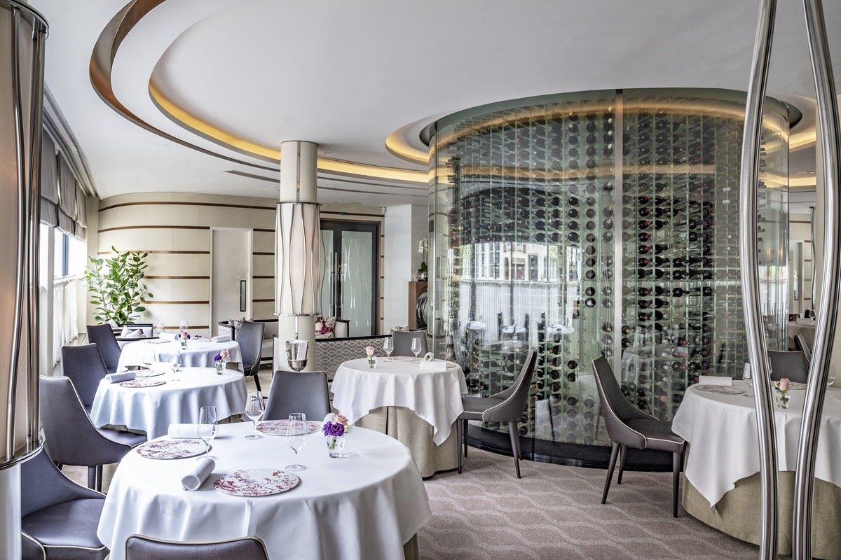 The interior of a fine dining restaurant with elegantly set tables, modern decor, and a large, cylindrical glass wine cellar.