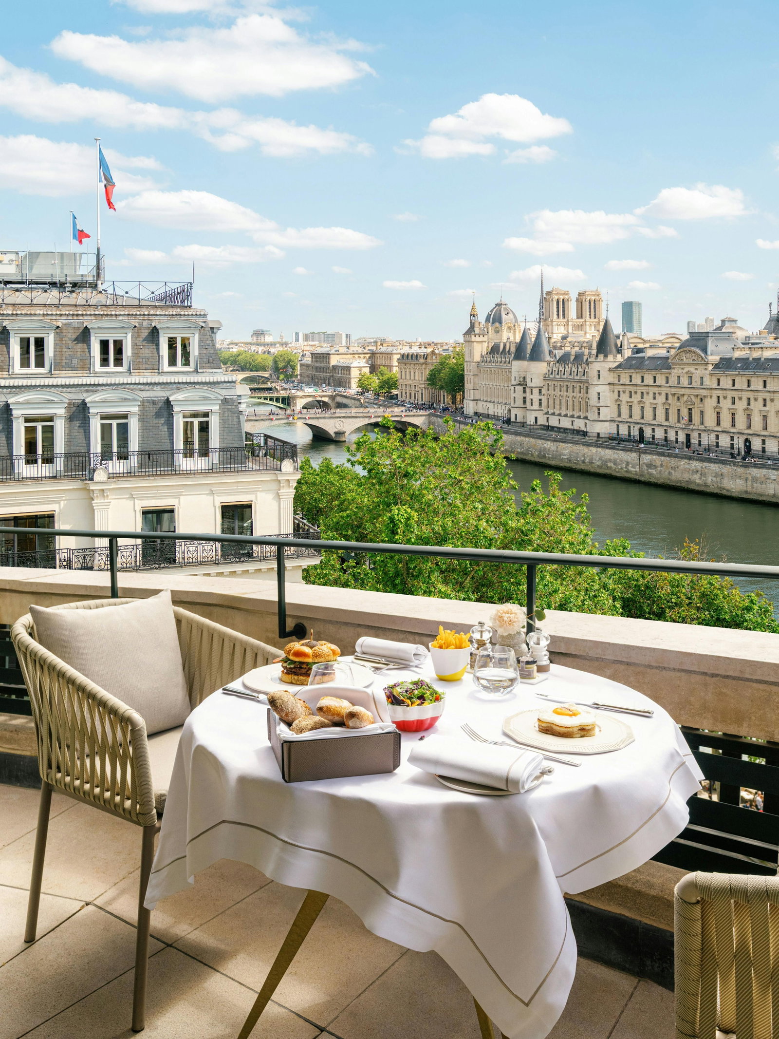 A balcony table set for a meal overlooking a river and city buildings, with a burger, fries, salad, and bread on the table.