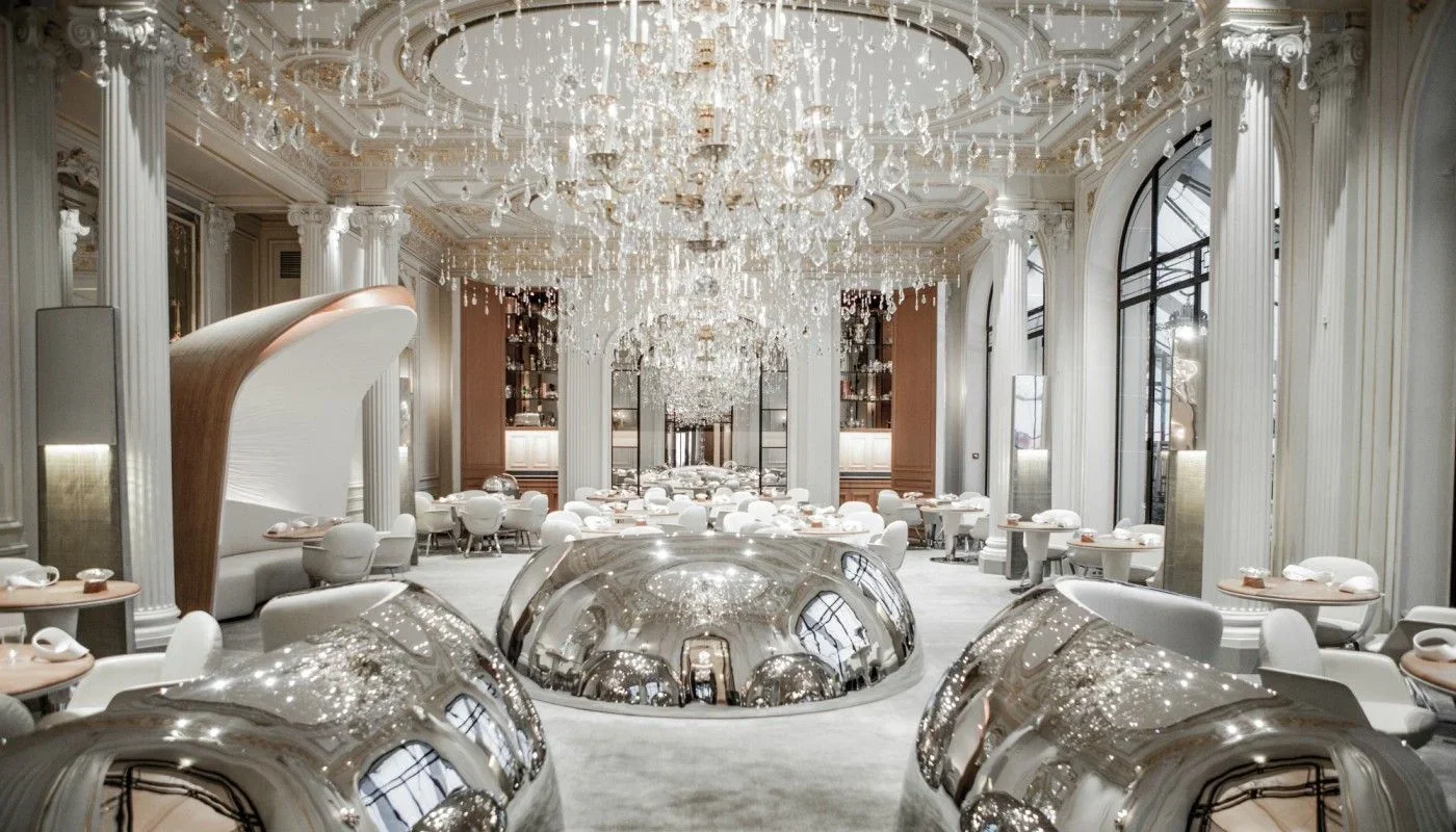 A luxurious fine dining restaurant with high ceilings, ornate chandeliers, and elegant white and gold decor. Several round tables with white chairs