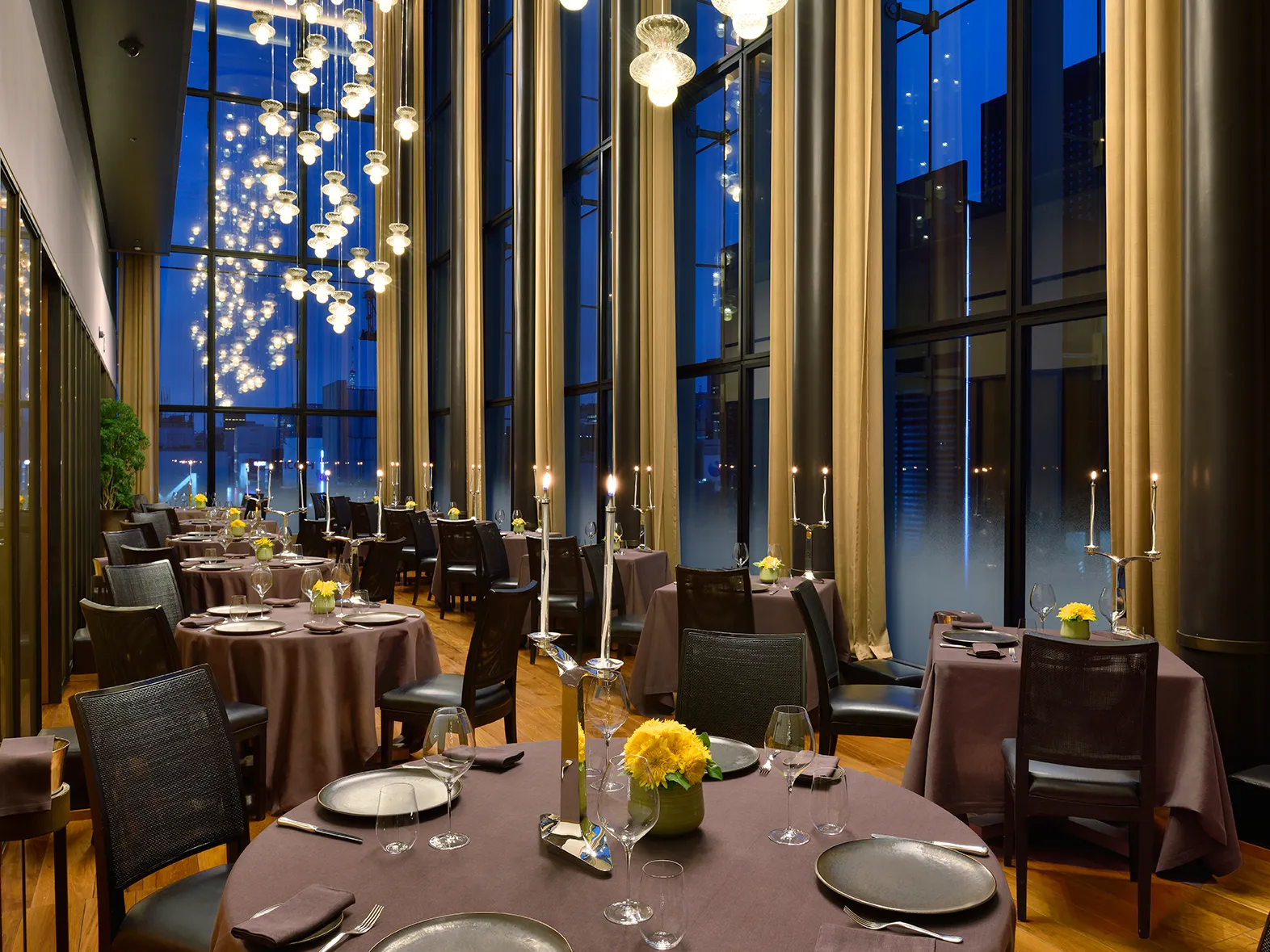 A fine dining restaurant in Tokyo with tall windows, long hanging lights, and tables set with dark tablecloths, plates, glasses, and yellow flower