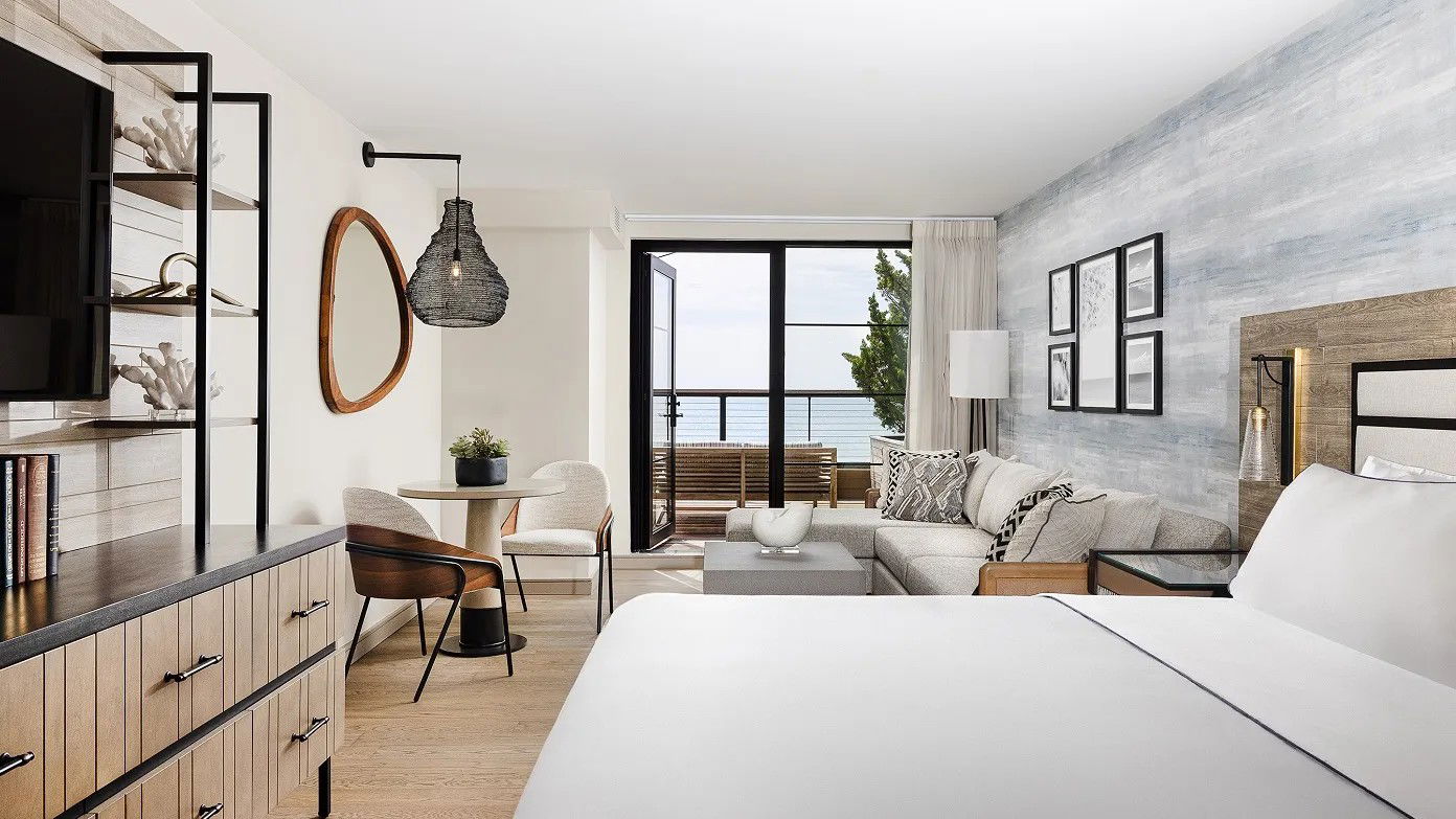 A modern hotel room with a king bed, a light wood dresser, a sectional sofa, a small dining table, and a balcony with an ocean view.