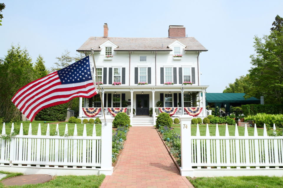 The front of the white Hedges Inn Hotel, featuring a large American flag, a picket fence, and a brick pathway leading to the entrance.