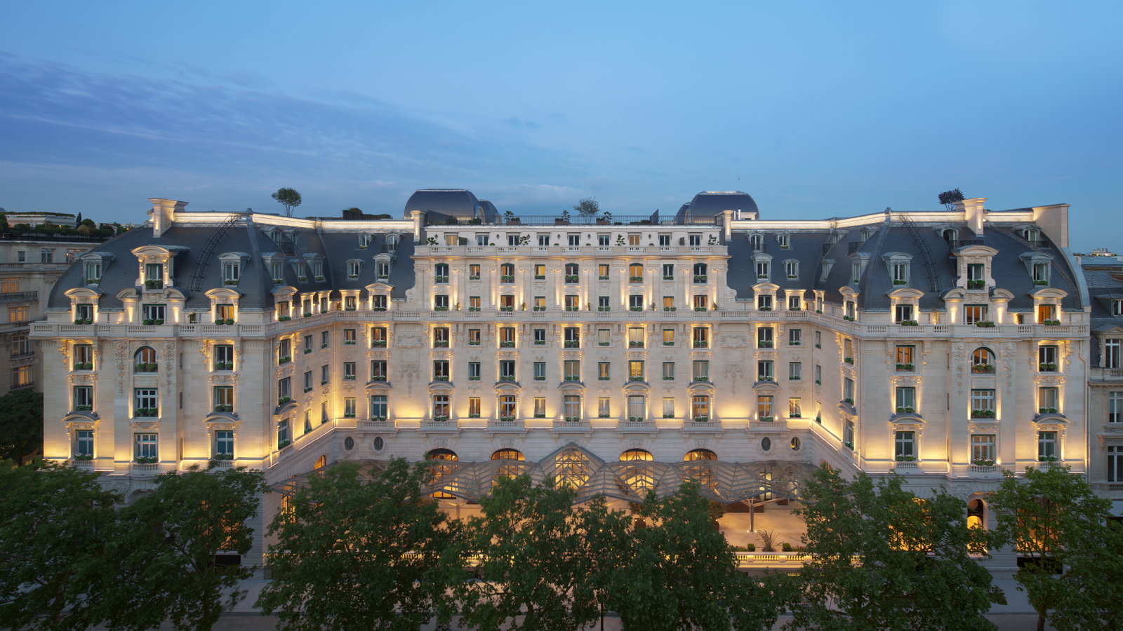 An illuminated luxury hotel building at dusk, featuring ornate architecture, many windows, and a dark, curved roof.