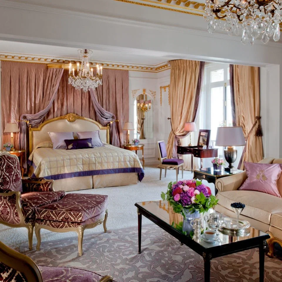 A luxurious hotel room with a large bed, elegant drapes, chandeliers, and a sitting area with plush furniture.