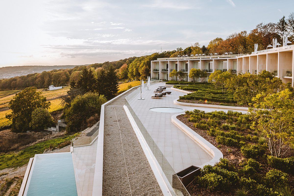 An aerial view of a modern luxury hotel with a long, curved building, multiple terraces, a swimming pool, and manicured gardens overlooking a valley